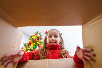 Surprised cute baby girl opens a Christmas gift. A small child opened a box next to a decorated...