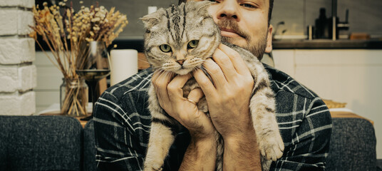 Scottish fold cat in hands of man with mustache wearing plaid shirt.