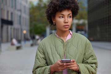 Horizontal shot of thoughtful woman wears jacket holds mobile phone looks away on promenade checks...