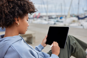 Outdoor shot of creative woman draws with stylus in digital tablet with mockup screen wears hoodie...