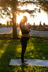 Side view of young woman expecting baby in black suit stands on rug barefoot and meditates on warm sunny summer day. Concept of sports and meditation activities