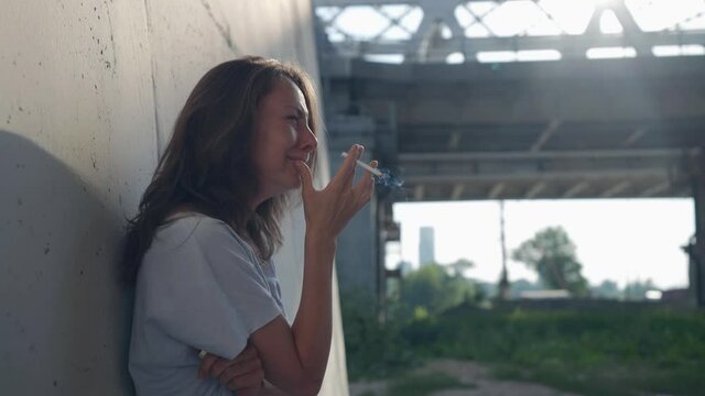 female portrait on the background of a railway bridge is out of focus. the woman leaned against the wall, clutched a cigarette with fingers, sobs and smokes