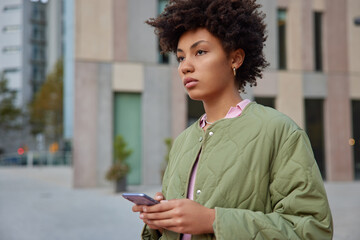 Sideways shot of curly haired young woman surfs mobile phone at street wears streetwear focused...