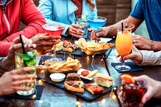 Group of friends enjoying appetizer drinking and eating in a bar - Close-up of hands of young people holding colorful cocktails in the happy hour time - Social gathering party time concept .