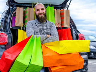 Happy young bald bearded man with car trunk full of colorful bags after a day of Christmas shopping...