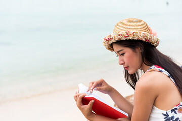 close up a young brunette relaxing on the beach, reading a book
Brunette girl in a dress with a hat and a jute bag sitting on a white beach and reading a book