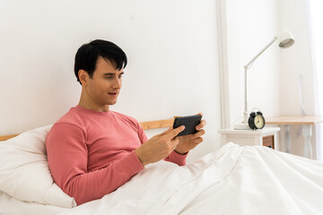 Smiling male awake lying in comfortable white bed checking mail on smartphone, happy biracial man wake up in morning using smartphone get pleasant message or text, Hangover,lazy and sleepless concept.