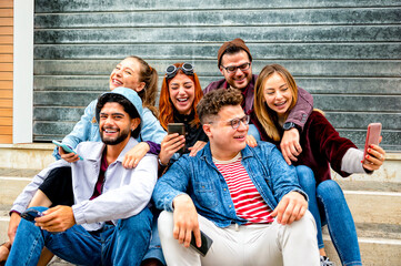 Happy multiracial friends sitting on the stairs laugh while chatting with smart phones - Students...