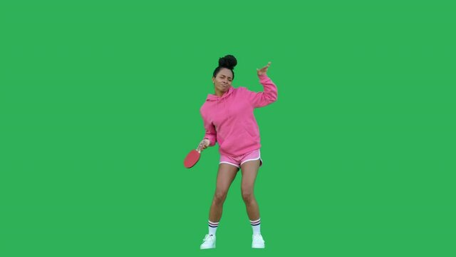 Young woman with black curly hair dancing over green background. African american girl table tennis player wearing pink hoodie and shorts having fun with racket. Chroma key. Copy space