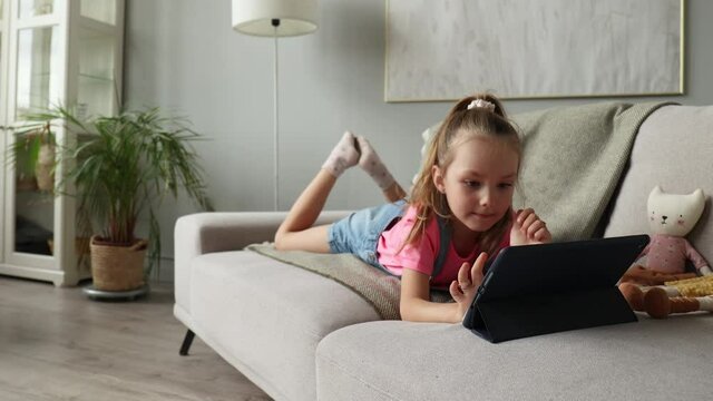 Smiling little girl lying on sofa playing online games, web surfing information, using funny applications on tablet.