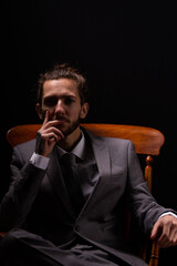 A young businessman in a smart gray suit sitting in a wooden arm chair with moody and atmospheric lighting