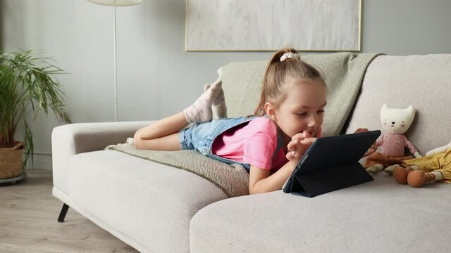 Smiling little girl lying on sofa playing online games, web surfing information, using funny applications on tablet.