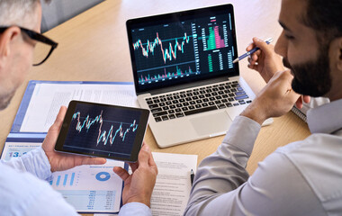 Fototapeta na wymiar Two diverse traders brokers stock exchange market investors discussing crypto trading charts growth using digital tablet and laptop analyzing financial risks, investment profit, global rates forecast.