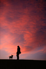 Obraz premium Silhouette of a woman with her dog standing against sunset sky with colorful clouds.