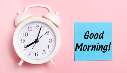 On a light pink background, a white alarm clock and a blue sheet of paper with the text GOOD MORNING