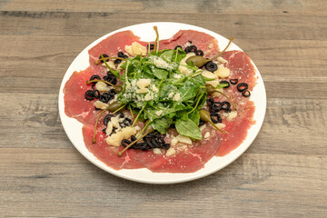 Delicious Italian recipe for carpaccio with parmesan cheese, slices of black olives, olive oil, arugula and basil, beef and capers in vinegar