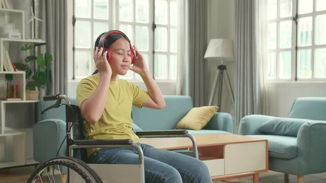 Asian Kid Girl Sitting In A Wheelchair Listening To Music With Headphones And Dancing In Living Room
