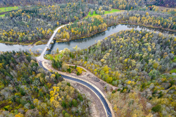 Aerial view from drone of concrete road intersecting big blue river among autumn pine, foliage forests in yellow green gold colors. Treetops in cloudy day and empty highway in fall season