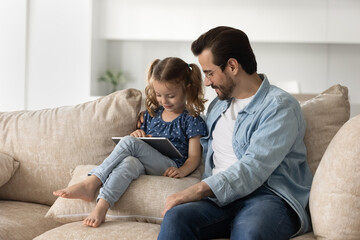 Caring smiling young father in eyeglasses watching happy little preschool kid daughter playing games on digital tablet, resting together on comfortable sofa, parental control, tech addiction concept.