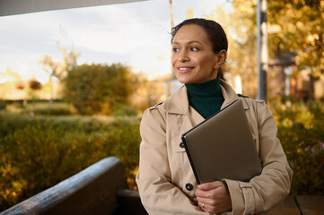 Confident middle aged beautiful woman, freelance, business person working on laptop, standing in the outdoor cafe on at beautiful autumnal sunset in the oak grove park. Start up, business concept