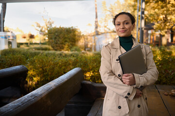 Confident portrait of attractive African business woman in casual beige trench coat and green pullover looking at the camera, posing with crossed arms holding laptop against autumn nature background