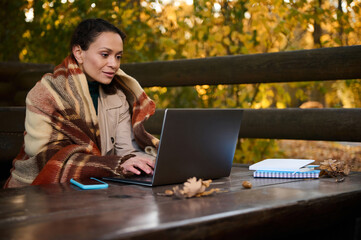Freelancer middle aged woman, developer wrapped in warm woolen blanket, types on laptop keyboard, works project, plans startup, sits at wooden table in country house, works remotely on cool autumn day