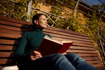 Fototapeta premium Relaxed pretty woman in casual denim and green pullover reading a book sitting on a wooden bench enjoying a warm sunny autumn weekend in country park, away from the hustle and bustle of the city