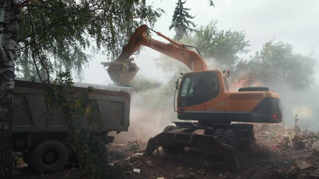 Demolition of building, excavator at work, bucket loads debris and rubble with lot of dust into truck for removal.