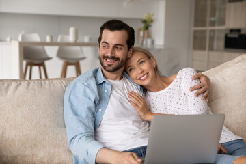 Happy bonding dreamy young family couple sitting on cozy sofa with computer on laps, thinking on received email with good news, visualizing future together in modern living room, feeling relaxed.
