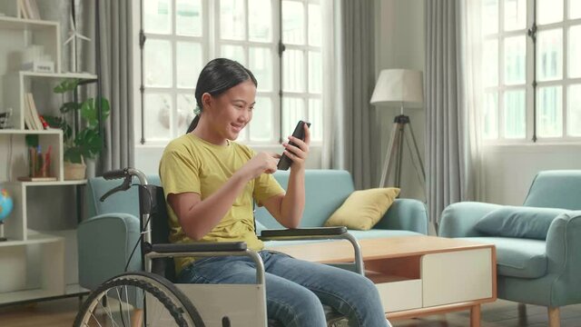 Happy Asian Kid Girl Sitting In A Wheelchair While Using Smart Phone And Celebrating At Home
