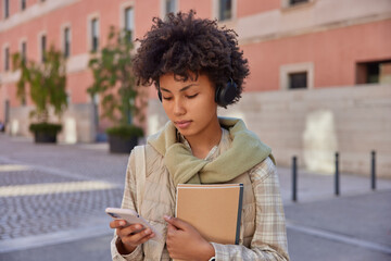 Photo of female student returns back to university after quarantine carries notepads focused at smartphone screen reads received notification listens audio lecture via headphones poses outside