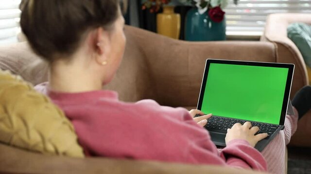 Young woman, girl relaxing at home on the couch and using laptop computer. Back view of girl watching movie on laptop at home. Green screen Over the shoulder shot of young woman working on modern lapt