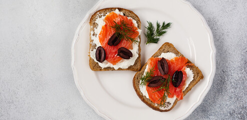 Sandwiches with smoked pink salmon olives, kalamata, microgreens and cream cheese on gray ceramic plate and trendy concrete background. Traditional Scandinavian toast. Top view
