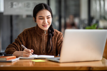 Fototapeta premium Young attractive university student using a laptop computer, studying online at home. Cheerful caucasian asian woman writes notes, planning working process, sitting at home. Exam preparation.