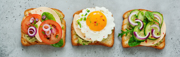 Three different toast sandwiches with guacamole sauce, fried egg, fresh cucumber, cheese and tomato with chard, arugula, red onion on gray concrete background. Selective focus