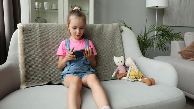 Smiling little girl sitting on sofa playing online games, web surfing information, using funny applications on smart phone.