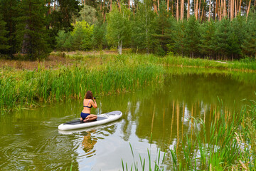 A woman drives on the Sup Board through a narrow canal surrounded by dense grass. Active weekend vacations wild nature outdoor. The woman is sitting on the lap in a bathing suit.