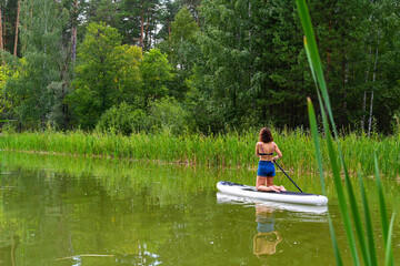 A woman drives on the Sup Board through a narrow canal surrounded by dense grass. Active weekend vacations wild nature outdoor. The woman is standing on the lap in a jeans.