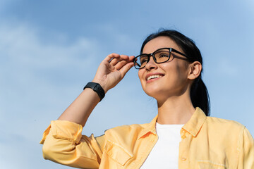 Looking at my perspectives. Low angle view of the satisfied woman touching her glasses and looking at the distance while walking at the street during the sunny day. Stock photo