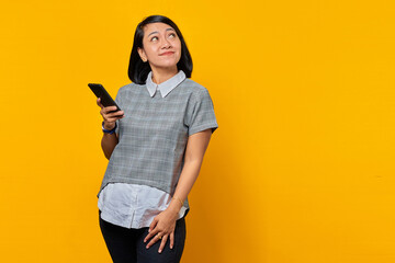 Beautiful smiling Asian woman holding smartphone and looking up over yellow background