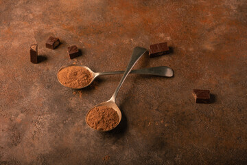 Cocoa powder in two deep vintage spoons, broken pieces of chocolate around, on a textured rusty background.