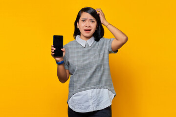 Portrait of beautiful asian woman holding smartphone touching her head and expressing concern on yellow background