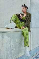 Vertical shot of pensive sad teenage girl dressed in fashionable apparel white boots sits against...