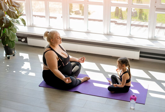 Young Obese Woman with cute little skinny daughter Doing Yoga, Meditation in lotus position at home in a room with a window on a sunny day.Mothers, baby sitting in pose position yoga pranayama balance