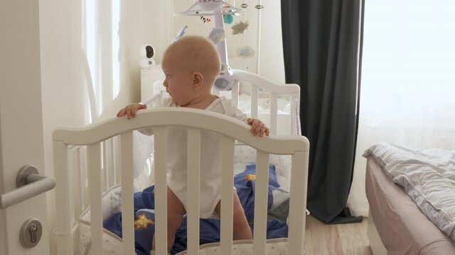 Happy cute adorable infant baby boy standing holding hands on crib playing in white rocking cradle at home. High quality 4k footage