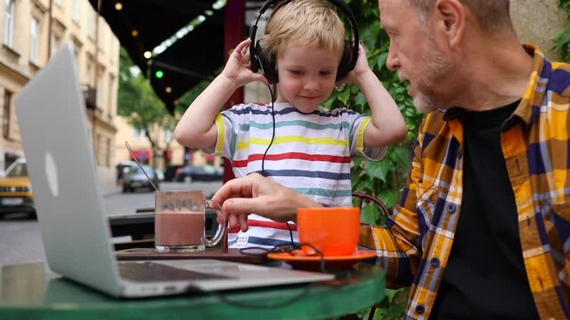 Smiling little boy using earphones and laptop with grandfather drinking cacao outside in cafe enjoying beautiful sunny day. Freelance urban lifestyle. Happy adult bearded man talking with grandson. 