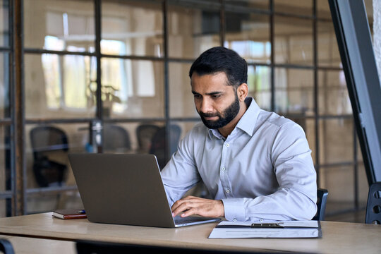 Serious Indian Business Man Employee Professional Manager Using Computer, Watching Webinar Working In Modern Office Doing Online Data Market Analysis Thinking Planning Tech Strategy Looking At Laptop.