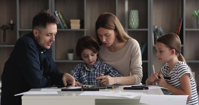 Caring young couple parents involved in creative domestic activity with joyful little children son and daughter, enjoying drawing together, sitting at table in modern living, early development concept