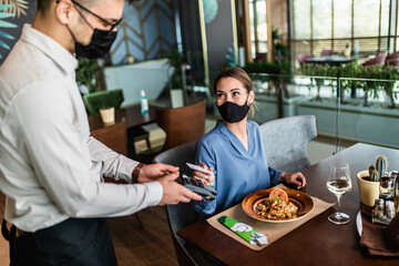 Beautiful young woman paying for her order with a credit card in a restaurant. She is wearing protective face mask as protection against Coronavirus or Covid-19 virus pandemic.