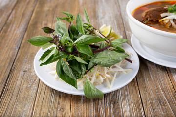A view of a plate of pho food accessories, featuring Thai basil, mung bean sprouts, lime wedge, hoisin sauce, and sriracha sauce.